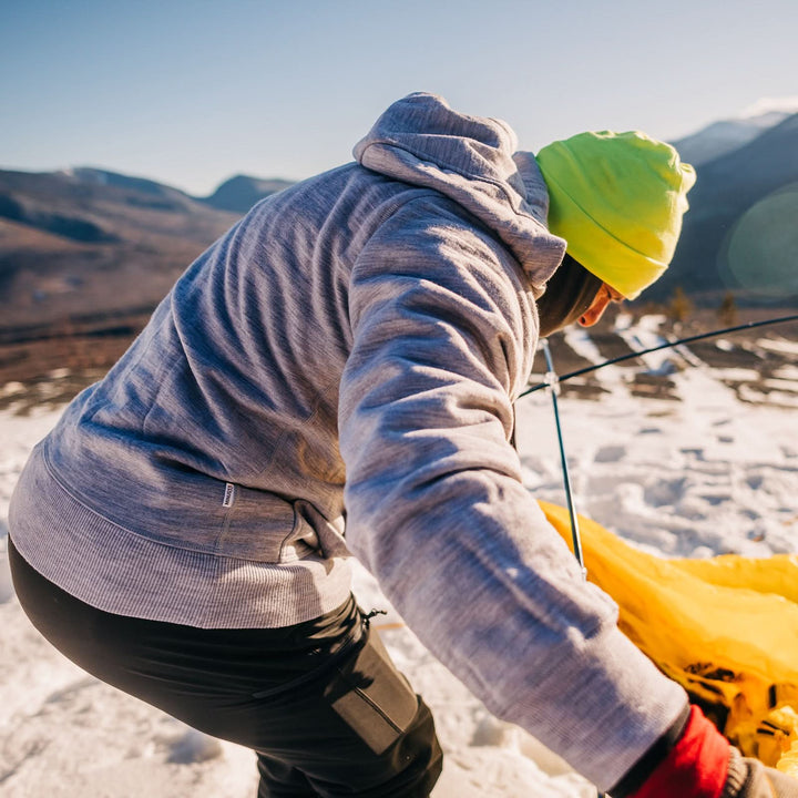A man sets up his tent on a snowy mountain. The man is modeling the heavyweight brushed wool Kodiak Fleece Hoodie, crafted with thumbholes and a drawstring to bundle you up for wicked cold days! #color_ash-gray