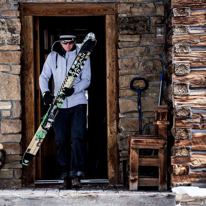 A man comes out of a cabin with skis in hand. The man is modeling the heavyweight brushed wool Kodiak Fleece Hoodie, crafted with thumbholes and a drawstring to bundle you up for wicked cold days! #color_ash-gray