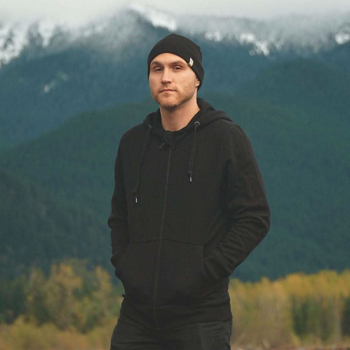 A man stands in front of mountains wearing the heavyweight brushed wool Kodiak Fleece Hoodie, crafted with thumbholes and a drawstring to bundle you up for wicked cold days! #color_black