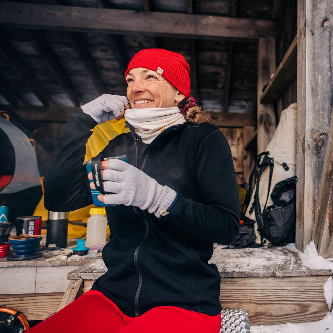 A woman sits on the step of her camp lodge to sip on a warm beverage from a thermos. The woman is wearing a Minus33 hat, neck gaiter, fingerless gloves, and a heavyweight merino wool full zip sweater. The sweater features a full length zipper, a two and a half inch collar, two zippered pockets, and thumbholes. #color_black