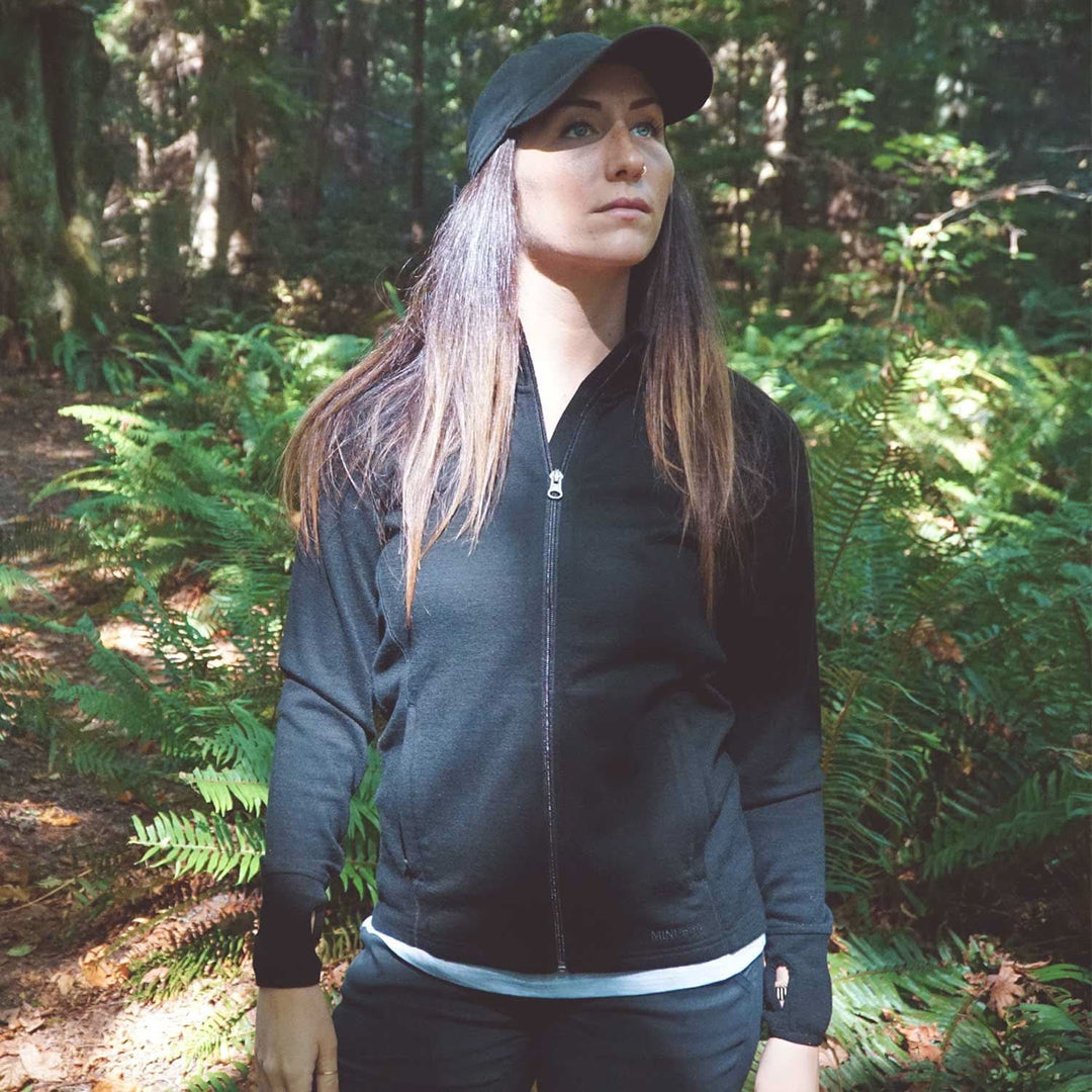 A woman stands in front of tall ferns surrounded by autumn leaves on the ground. Sunlight peeks through the trees. The woman is wearing a hundred percent merino wool heavyweight full zip sweater. This outer layer features a two and a half inch mock neck collar, two zippered pockets, and thumbholes. #color_black