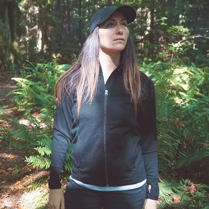 A woman stands in front of tall ferns surrounded by autumn leaves on the ground. Sunlight peeks through the trees. The woman is wearing a hundred percent merino wool heavyweight full zip sweater. This outer layer features a two and a half inch mock neck collar, two zippered pockets, and thumbholes. #color_black