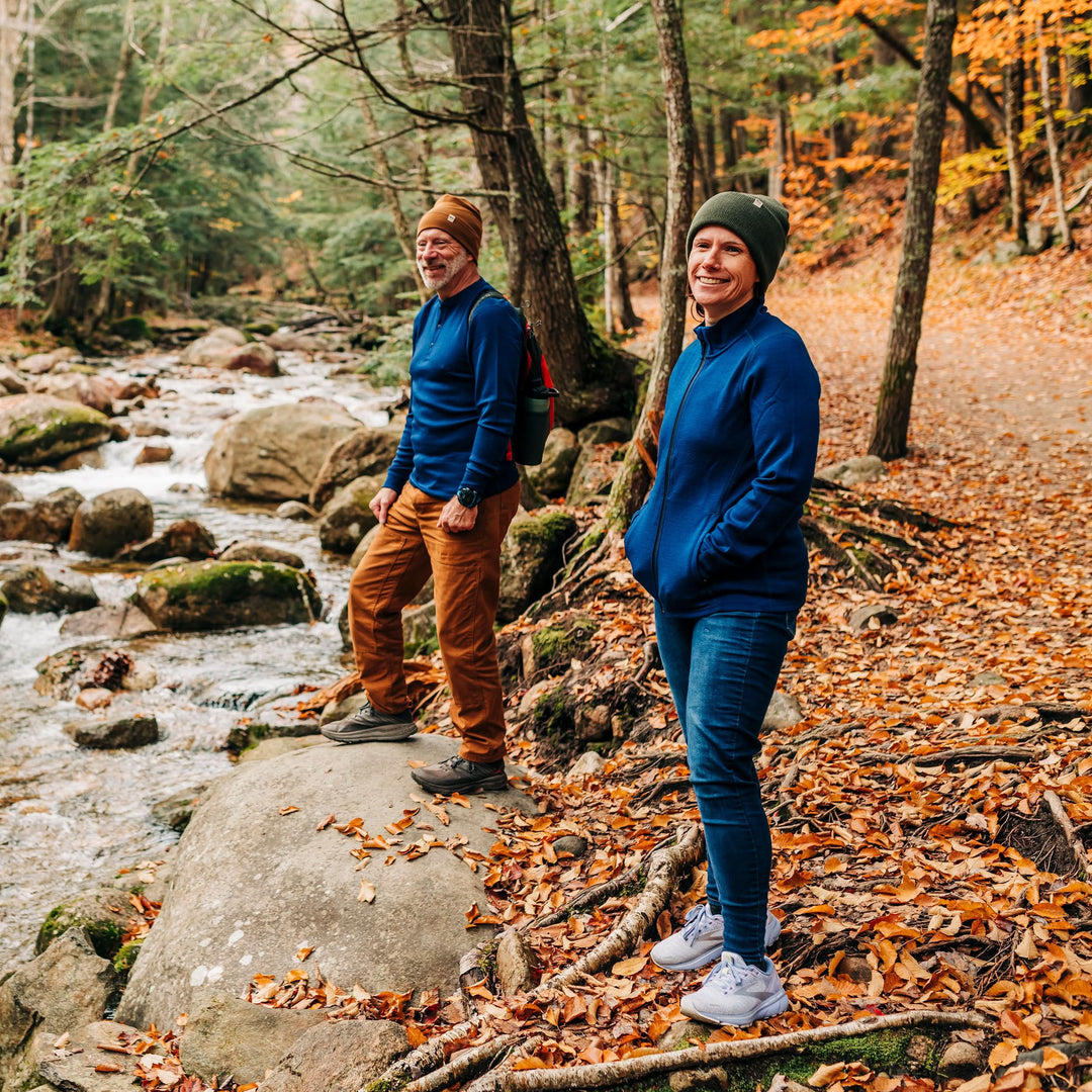 Two people stand on a trail covered in autumn leaves at the edge of a stony, flowing brook. The woman wears a merino wool hat and a hundred percent merino wool heavyweight full zip sweater. This outer layer features a two and a half inch mock neck collar, two zippered pockets, and thumbholes. #color_navy