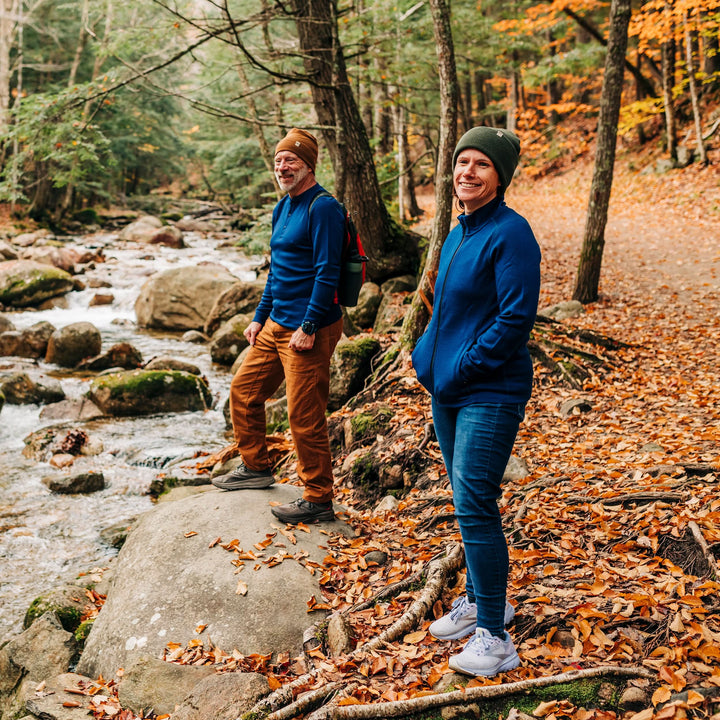 Two people stand on a trail covered in autumn leaves at the edge of a stony, flowing brook. The woman wears a merino wool hat and a hundred percent merino wool heavyweight full zip sweater. This outer layer features a two and a half inch mock neck collar, two zippered pockets, and thumbholes. #color_navy