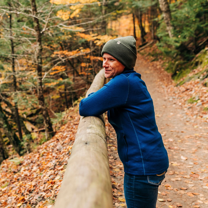 A woman stands on a walking trail in autumn, wearing her hundred percent merino wool heavyweight full zip sweater and a hat. This outer layer features a two and a half inch mock neck collar, two zippered pockets, and thumbholes. #color_navy