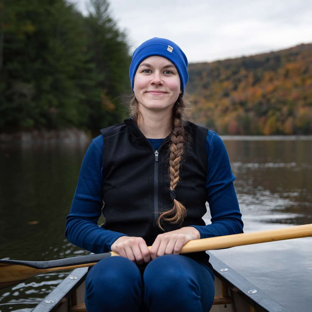 A woman sits in a canoe on open water in front of mountains and trees. The woman is wearing a lightweight hat, midweight base layer top and bottoms, and a hundred percent merino wool heavyweight full zip vest. This outer layer features a two and a half inch mock neck collar and two zippered pockets. #color_black