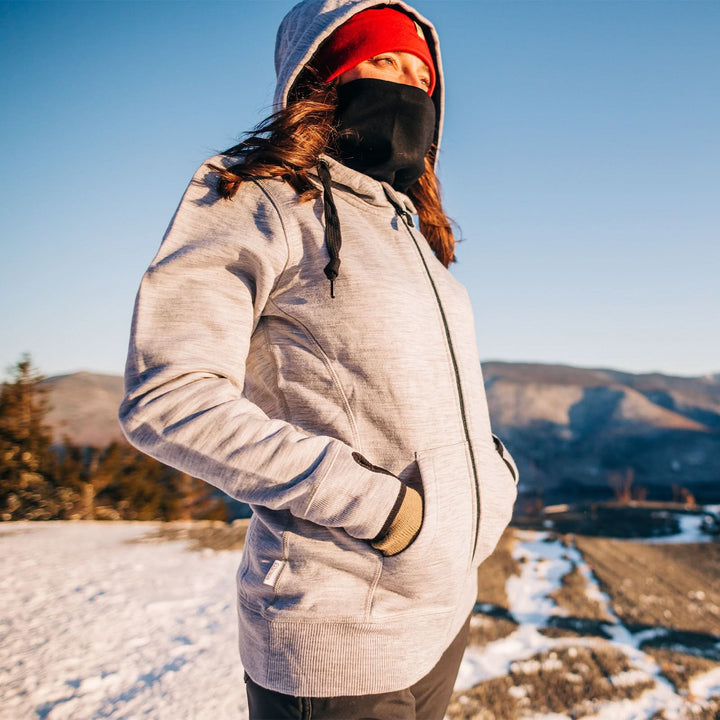 A woman stands in the snow looking out  toward the sun. The woman wears the heavyweight merino wool brushed fleece hoodie, a neck gaiter, and a lightweight beanie. The hoodie features a large hood with drawstrings, thumbholes, two pockets, and an interior stash pocket inside. #color_ash-gray