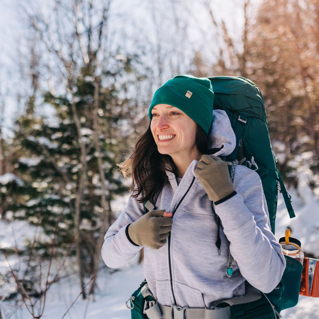 A woman hikes through the snowy forest wearing a backpack over the heavyweight merino wool brushed fleece hoodie. The hoodie features a large hood with drawstrings, thumbholes, two pockets, and an interior stash pocket inside. #color_ash-gray