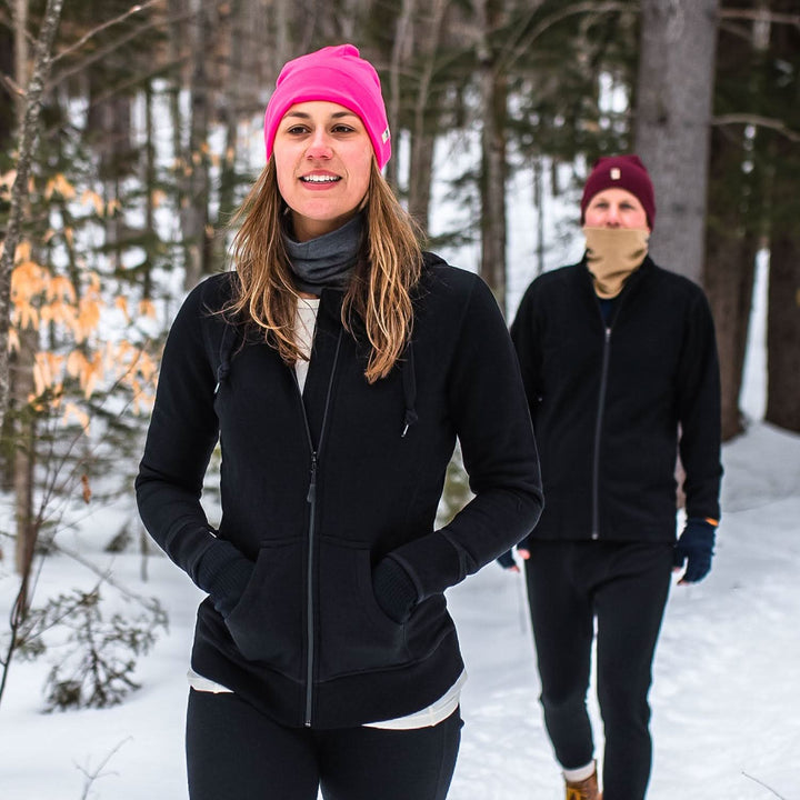 Two people walk through the snowy forest wearing their merino wool gear to keep warm. A woman models the heavyweight merino wool brushed fleece hoodie with a beanie and neck gaiter. The hoodie features a large hood with drawstrings, thumbholes, two pockets, and an interior stash pocket inside. #color_black