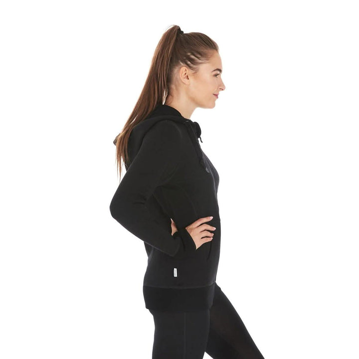 A woman models the heavyweight merino wool brushed fleece hoodie. The hoodie features a large hood with drawstrings, thumbholes, two pockets, and an interior stash pocket inside. #color_black