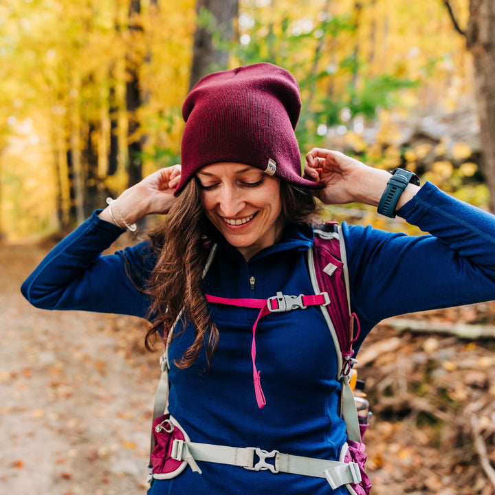 A woman on the trail models a heavyweight, super soft, one hundred percent merino wool beanie. This heavyweight beanie is the warmest hat that we offer, perfect for hiking through the White Mountains, or sitting at a bonfire in January. #color_burgundy