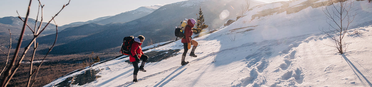 Wide panoramic view of two backpacked hikers walking through deep snow below frosted alpine trees.