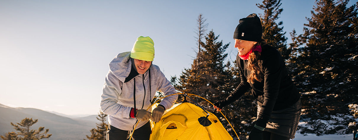 couple setting up a tent on top of a mountain in kodiak fleece hoodies in the winter