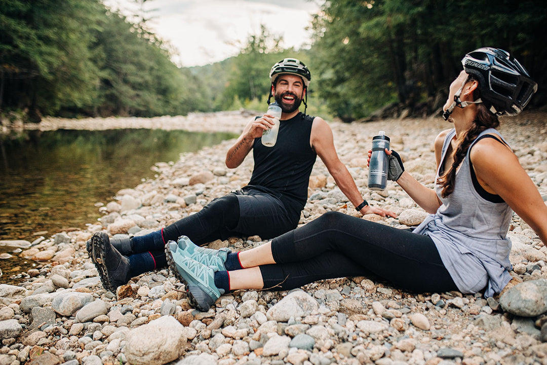 two people sitting next to a river wearing merino wool
