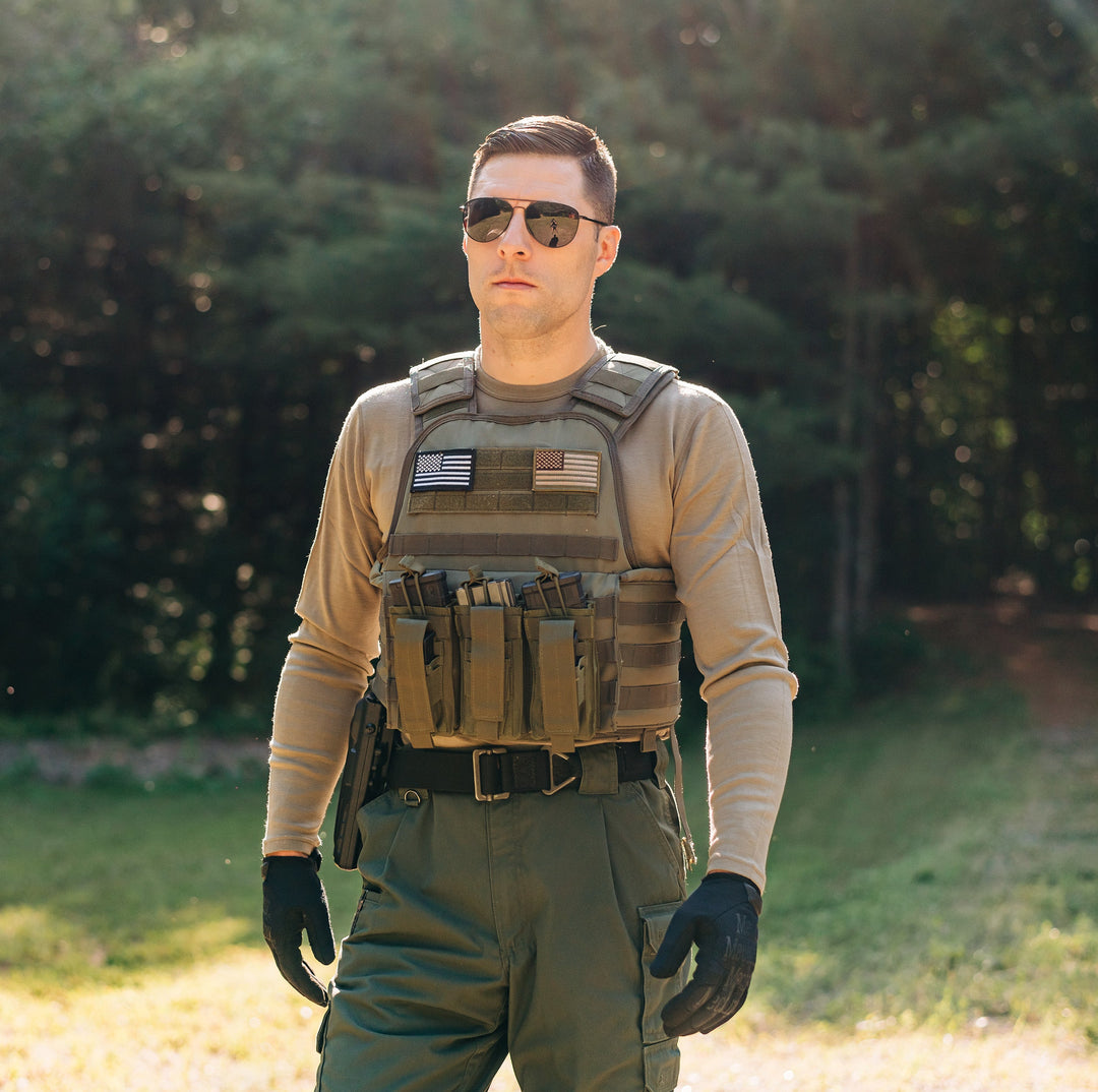 Man standing in a merino wool long sleeve shirt and a bullet proof vest. He has sunglasses on and black gloves and green pants