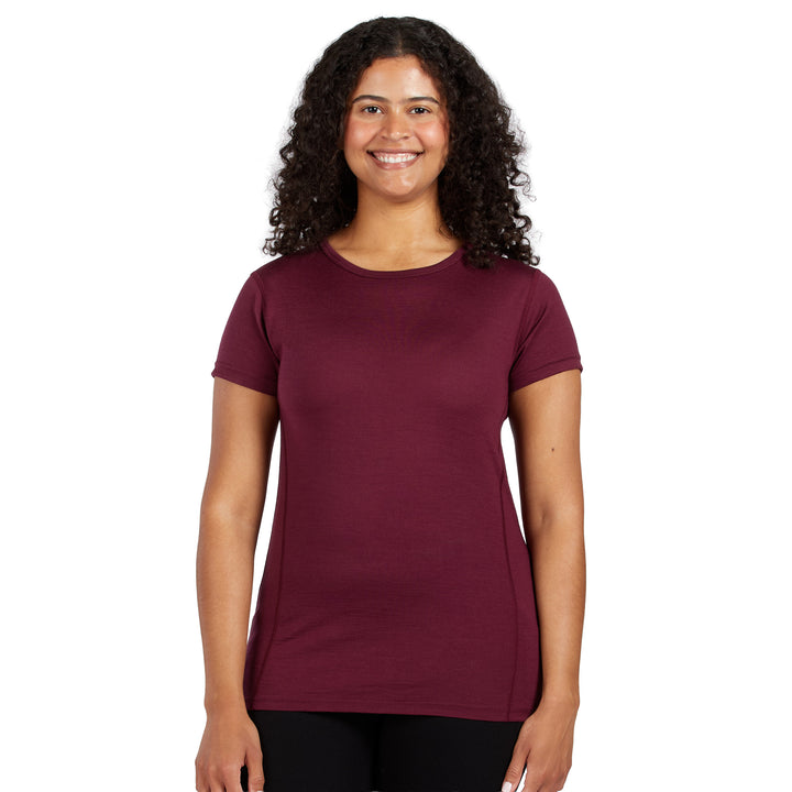 A smiling woman with curly dark hair wearing a short-sleeve 100% Merino wool crewneck t-shirt in the color burgundy, paired with black pants, against a solid white background. 