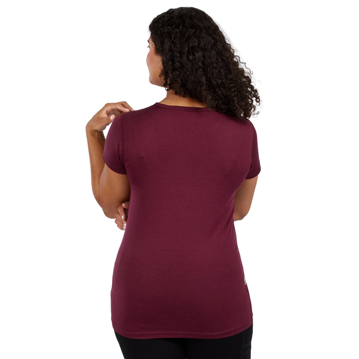 A woman with curly dark hair is shown from the back wearing a 100% Merino wool short-sleeve crewneck t-shirt in a burgundy color, paired with black pants, against a solid white background. 