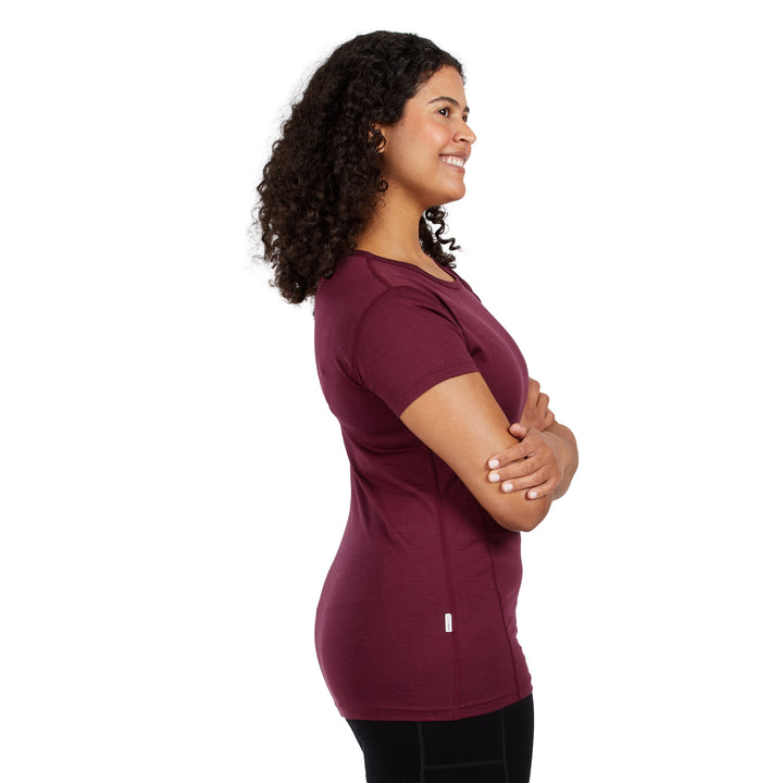 A woman with curly dark hair is shown in profile wearing a 100% Merino wool short-sleeve burgundy athletic top and black leggings against a plain white background. 