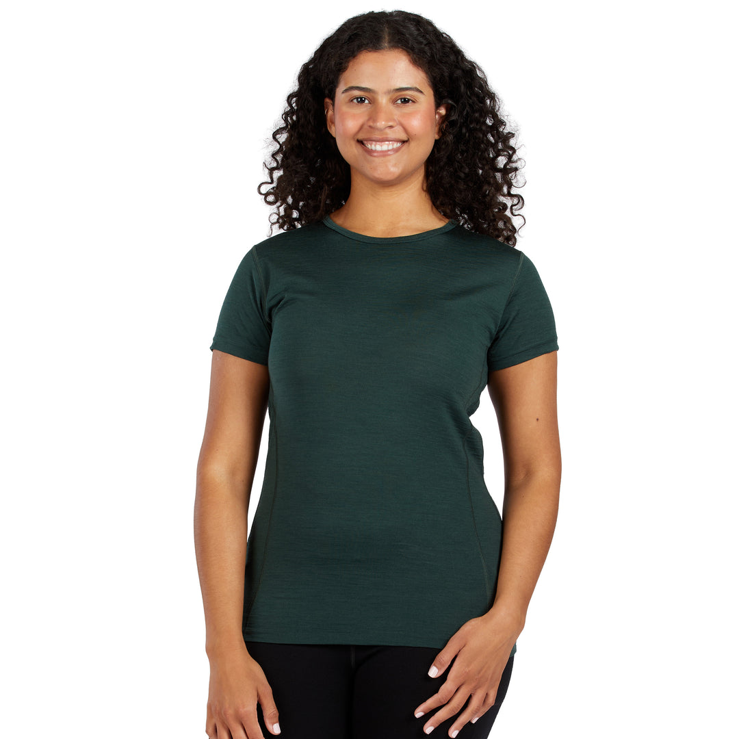 A smiling woman with curly dark hair wearing a dark green 100% merino wool short-sleeve crewneck t-shirt and black pants, standing against a plain white background. 