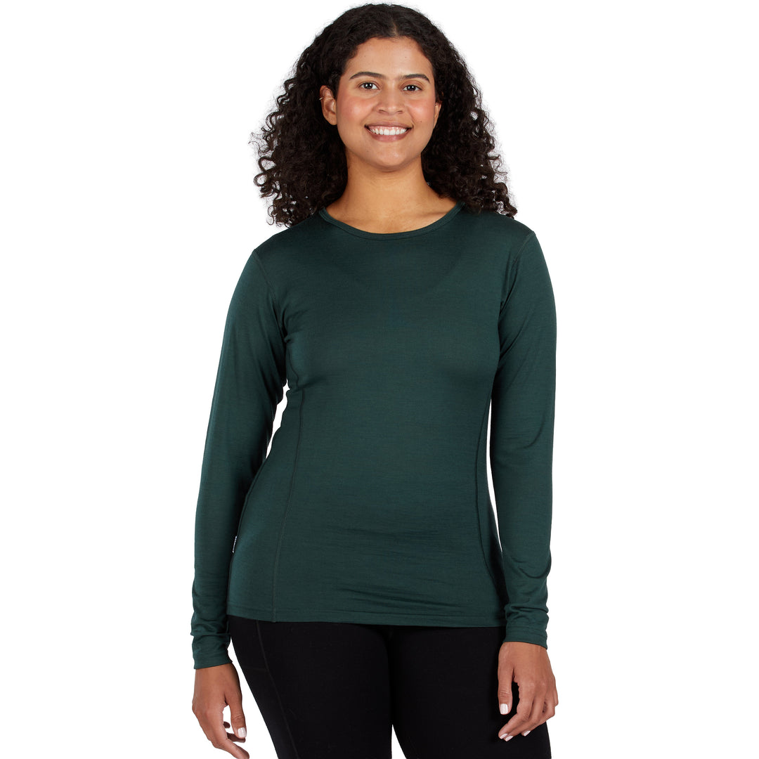 A smiling woman with curly dark hair wearing a dark green 100% Merino wool long-sleeve crewneck athletic top and black leggings, standing against a plain white background. 