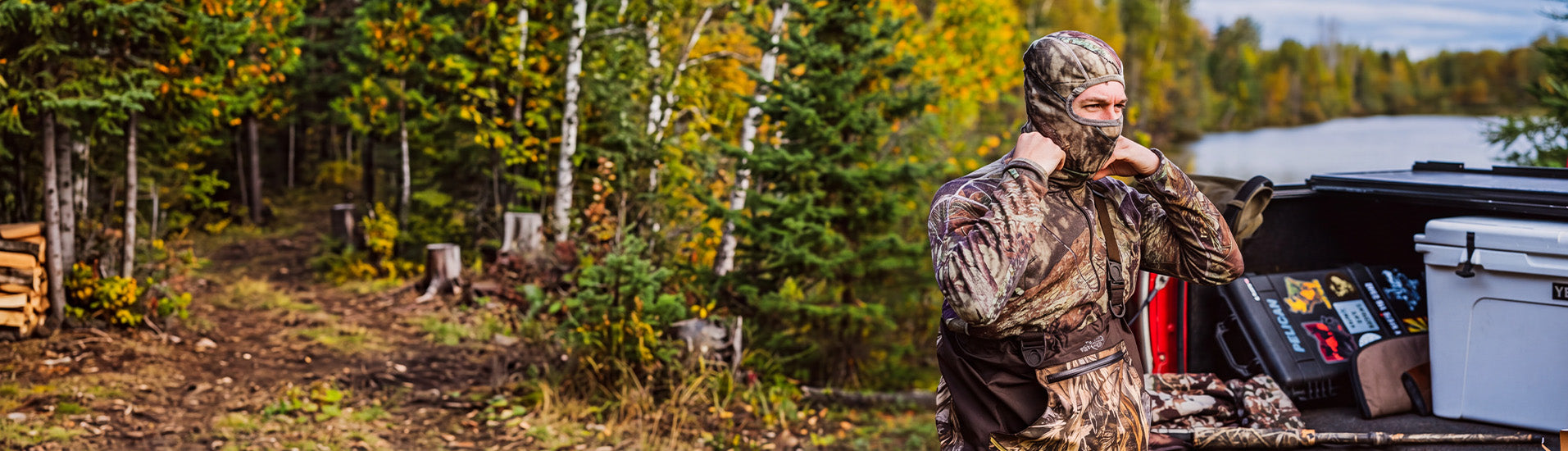A man in full merino wool camouflage hunting clothing stands by the open tailgate of a truck with a cooler and gear, with a forest and lake in the background.