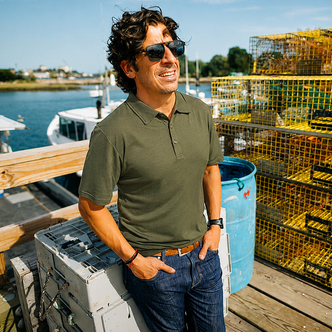Man standing on a dock with lobster traps and a boat in the background wearing a green merino wool polo 