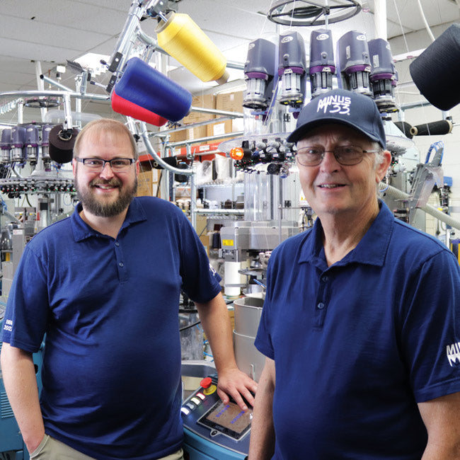 Two men in navy polo shirts standing among sock-making machines and colorful yarn spools in a factory.