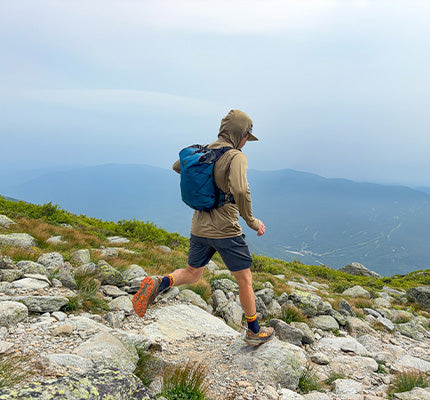 A person hiking on a rocky mountain trail wearing a beige hooded jacket, dark shorts, hiking shoes, and carrying a blue backpack, with distant blue mountain ridges under a cloudy sky.