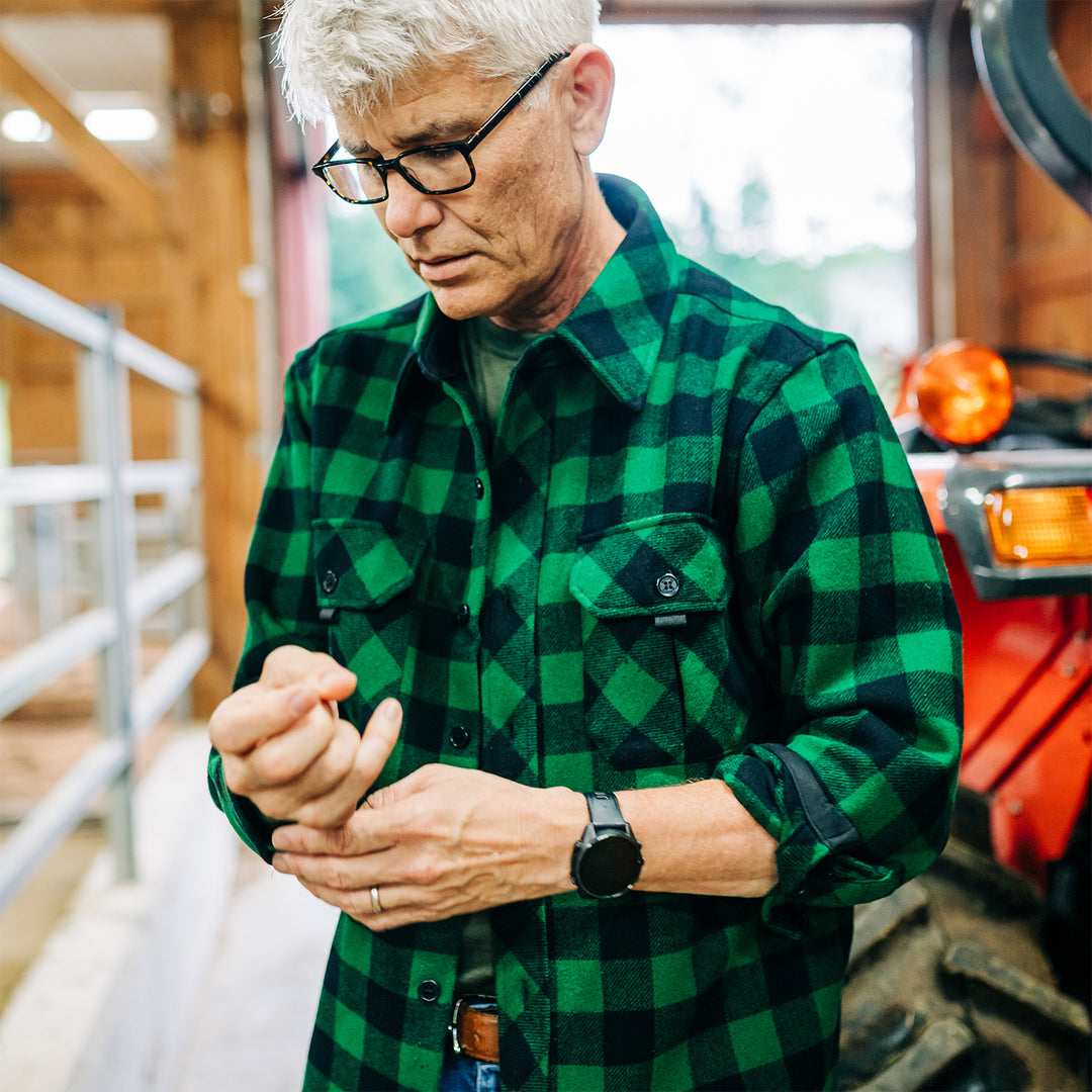 Man buttons green plaid heavyweight wool shirt in barn #color_green-plaid